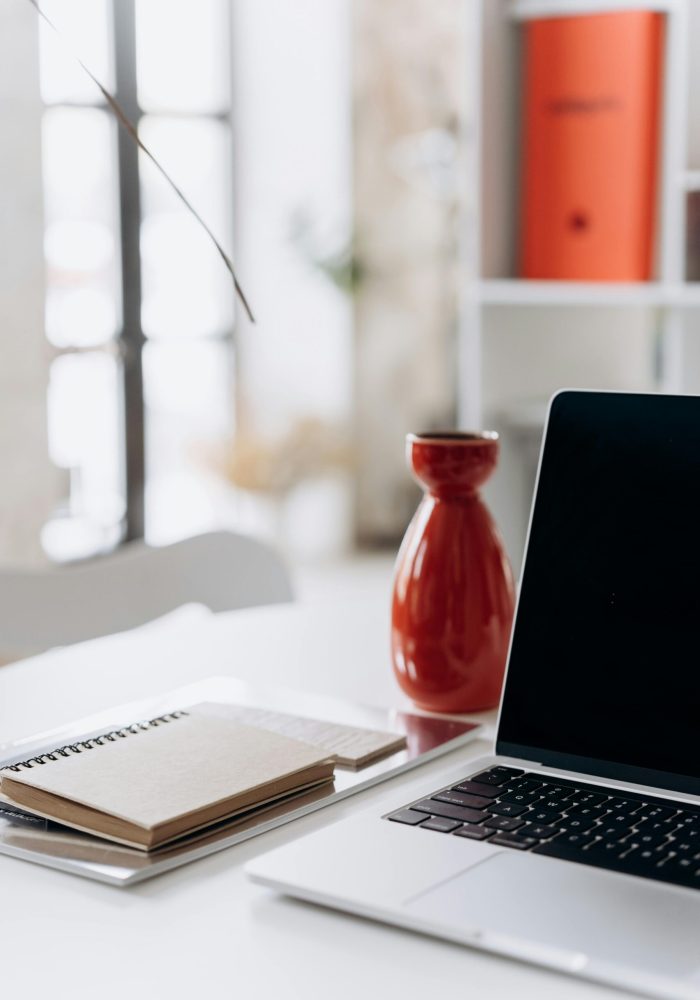 A minimalist home office setup featuring a laptop, notebook, and a red vase on a white desk.